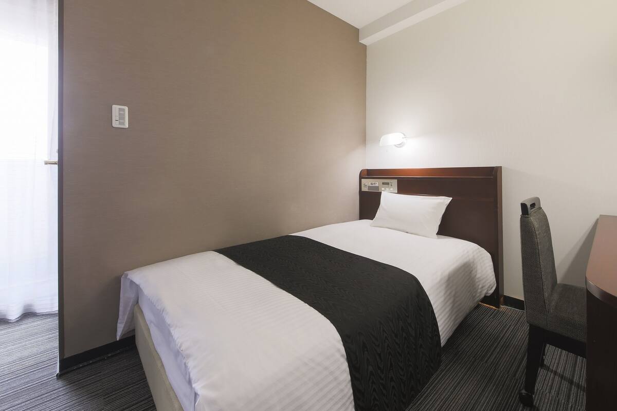 Habitación de hotel con una cama individual, una mesita de noche, una silla y una lámpara fijada a la pared.