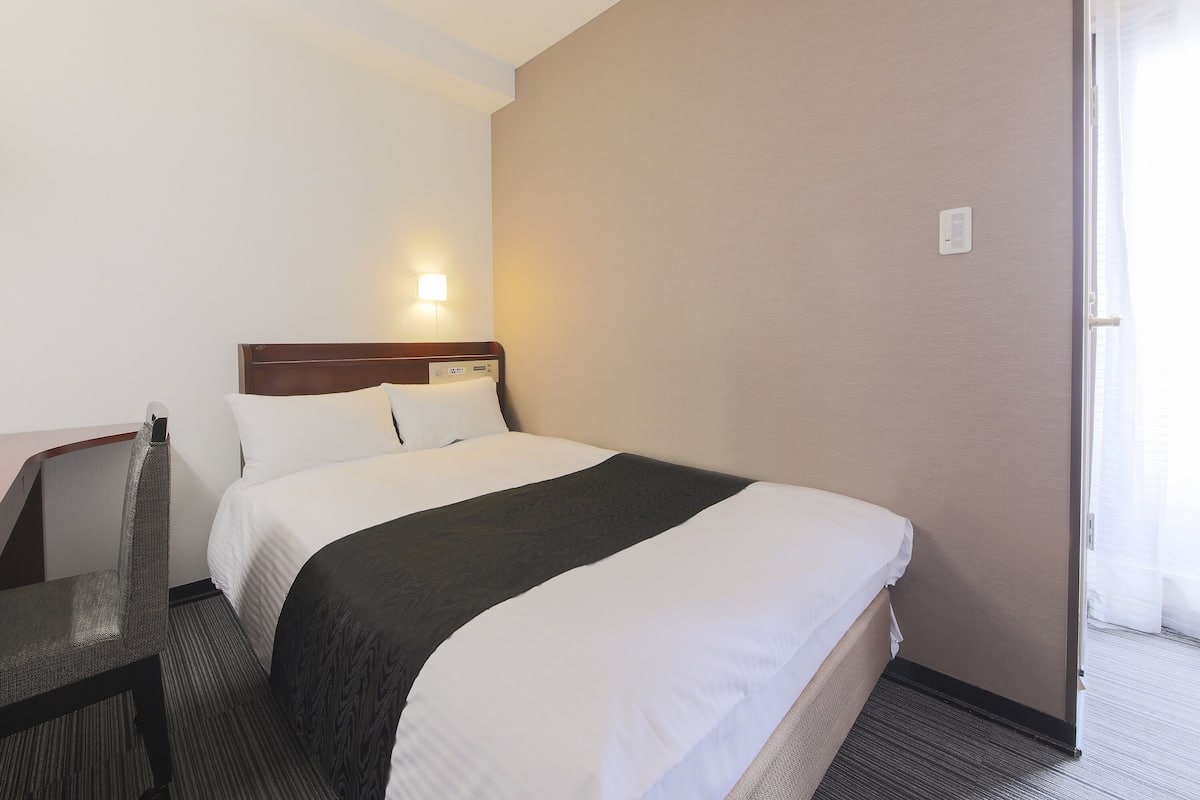 Una habitación de hotel con cama, una silla, una mesita de noche y una lámpara en la pared.