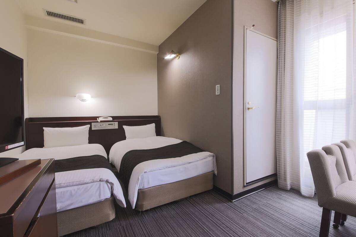 Habitación de hotel con dos camas, un escritorio, una silla, un televisor y una ventana con cortinas.