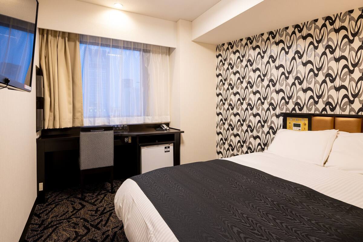 Ein Hotelzimmer mit einem Bett, einem Schreibtisch mit Computer, einer kleinen Kühlschrank und einem Fenster mit Vorhängen.