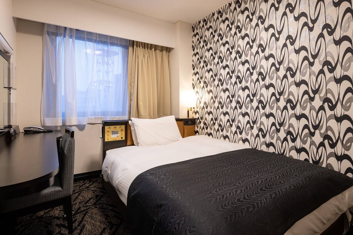 Ein Hotelzimmer mit Bett, Schreibtisch, Stuhl, Nachttisch, Lampe und einer Wand mit Muster.