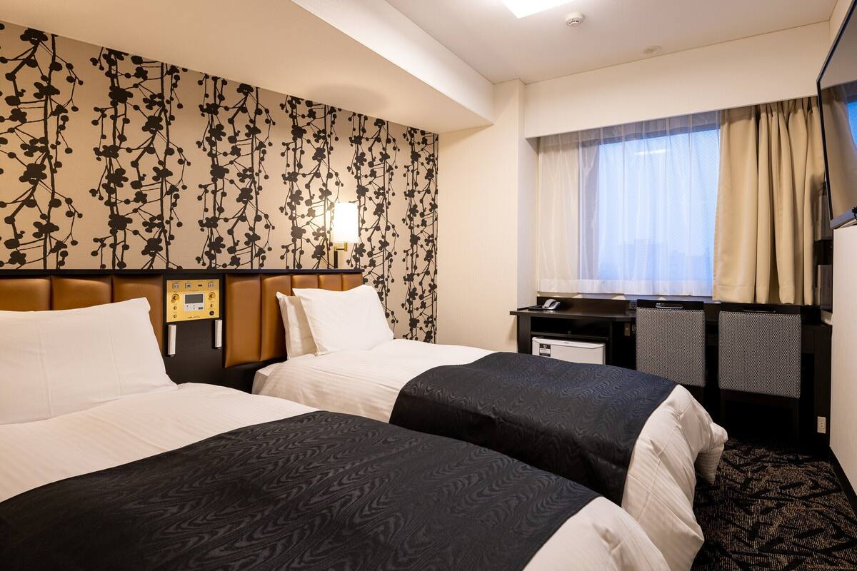 Ein Hotelzimmer mit zwei Betten, einem Schreibtisch, einem Stuhl, einem Fernseher und einem Fenster mit Vorhängen.