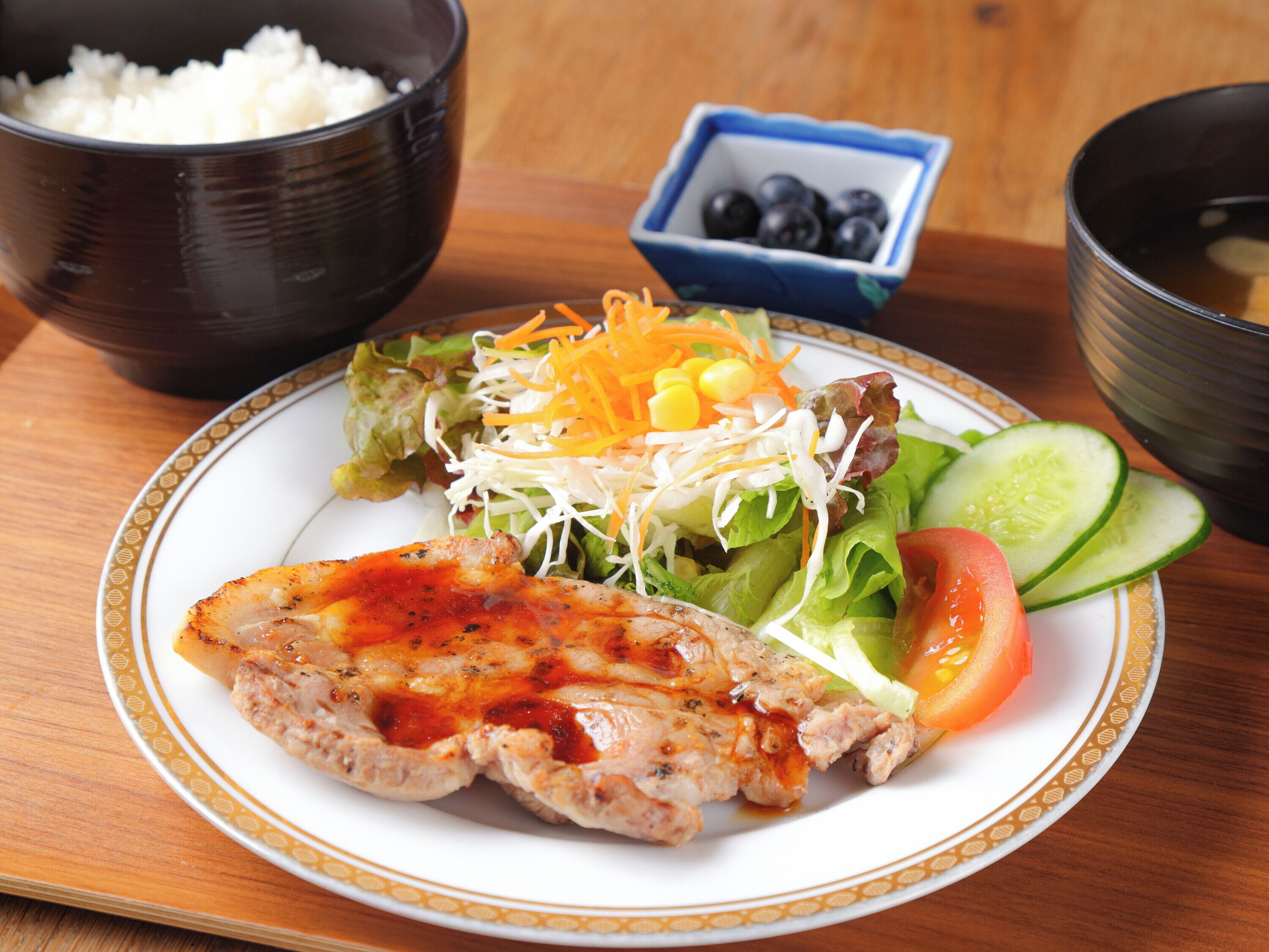 A plate with grilled chicken, salad, and vegetables on a table with a bowl of rice and a bowl of soup.