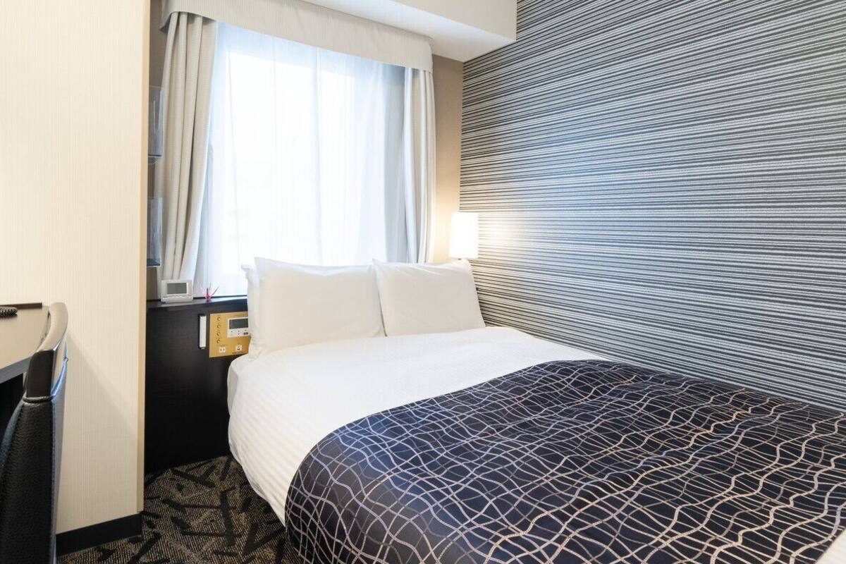 Ein Hotelzimmer mit Bett, Schreibtisch, Stuhl, Nachttisch, Lampe und einem Fenster mit Vorhängen.