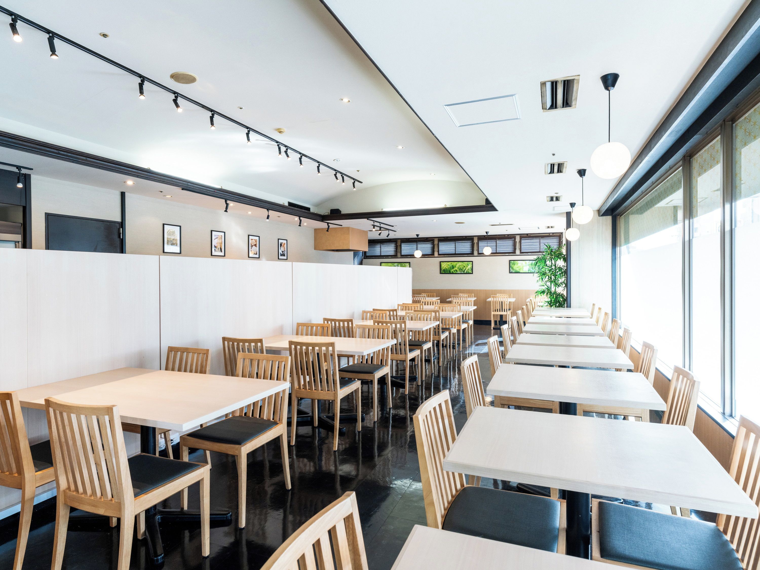A modern restaurant interior with multiple tables and chairs, a high ceiling with track lighting, and large windows allowing natural light.
