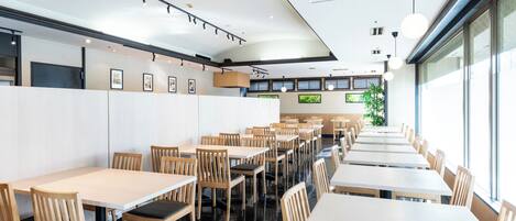 A modern restaurant interior with multiple tables and chairs, a high ceiling with track lighting, and large windows allowing natural light.