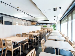 Un intérieur de restaurant moderne, avec de nombreuses tables et chaises, un plafond haut doté de luminaires sur rails et de grandes fenêtres laissant entrer la lumière naturelle.