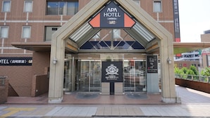 De ingang van APA HOTEL, met een modern ontwerp, voorzien van een glazen deur en een opvallend bord boven de deuren.
