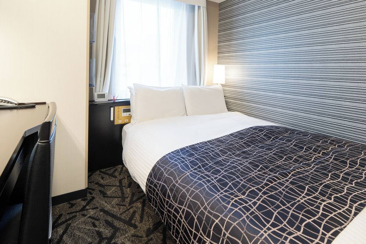 Ein Hotelzimmer mit einem Bett, einem Schreibtisch mit Telefon, einem Fenster mit Vorhängen und einer Wand mit Streifenmuster.