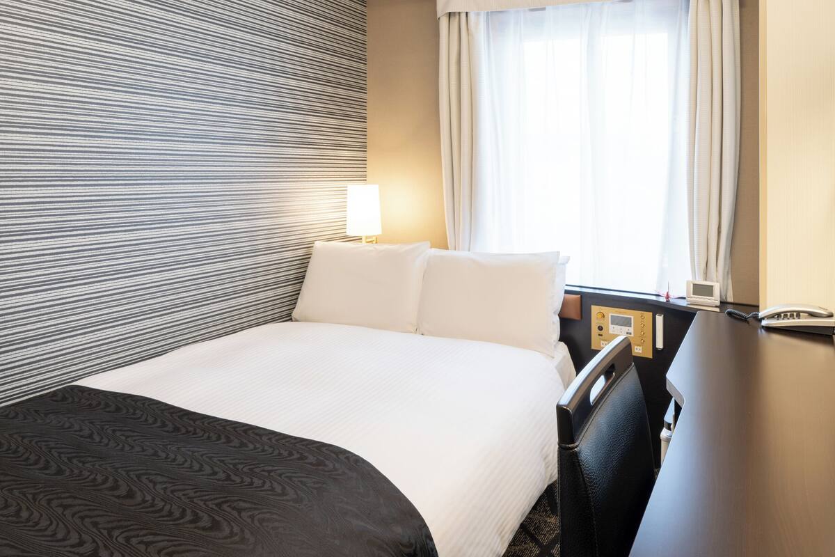 Une chambre d’hôtel avec un lit, un bureau, une lampe, un téléphone et une fenêtre avec des rideaux.