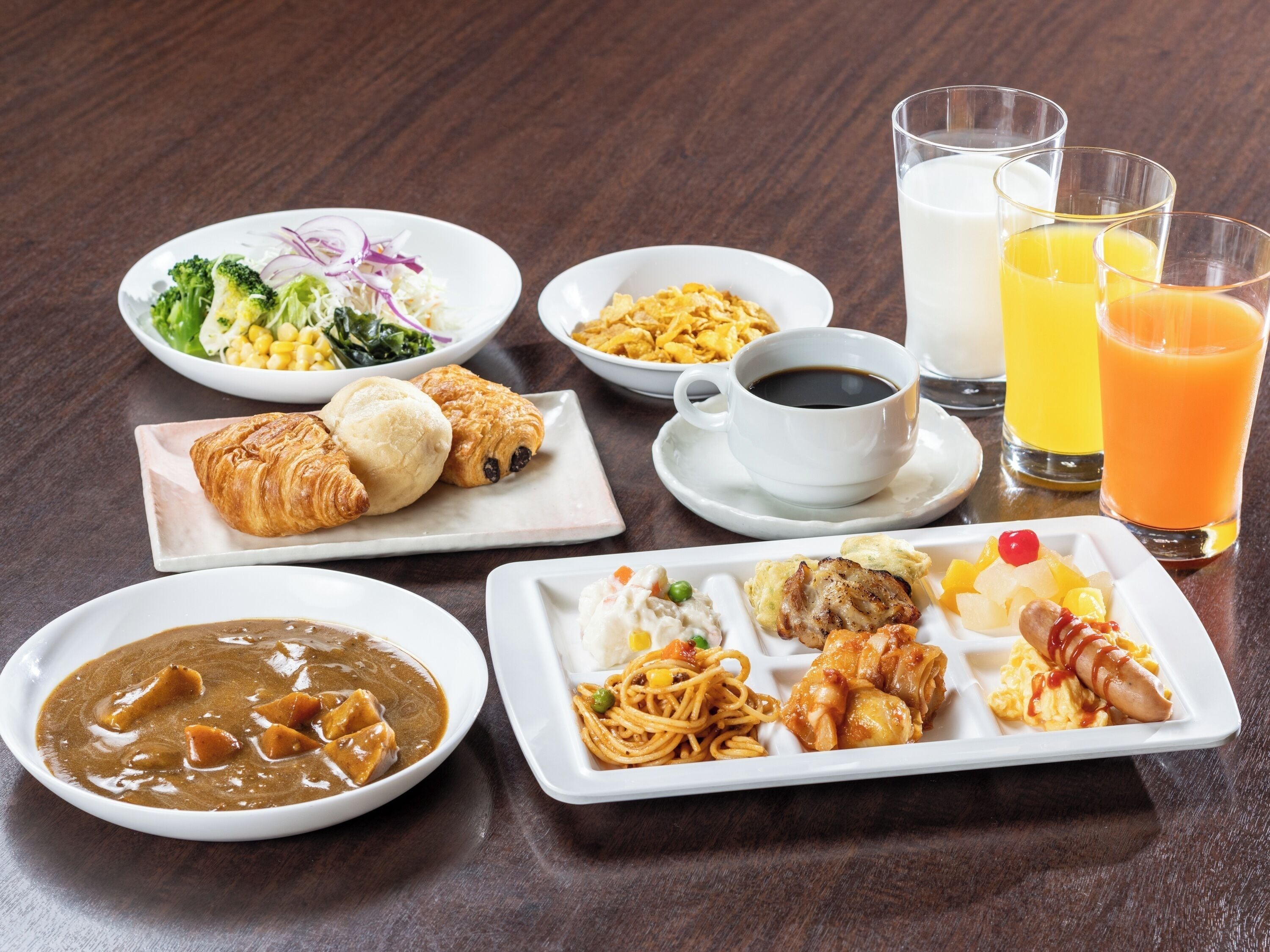 A variety of breakfast dishes including croissants, a bowl of soup, and a plate of noodles. There are also glasses of milk and orange juice.