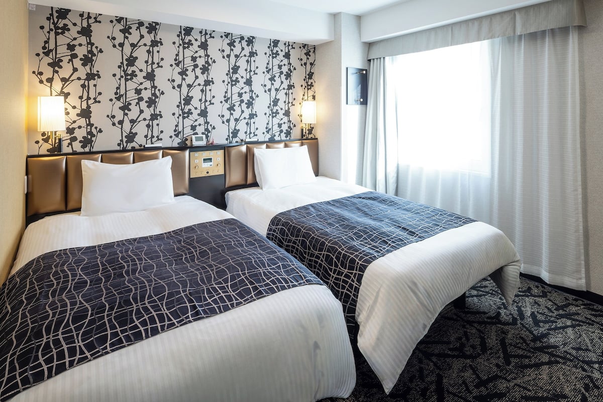 Ein Hotelzimmer mit zwei Betten, einer wandmontierten Steuerung, einem Fenster mit Vorhängen und einem gemusterten Teppich.