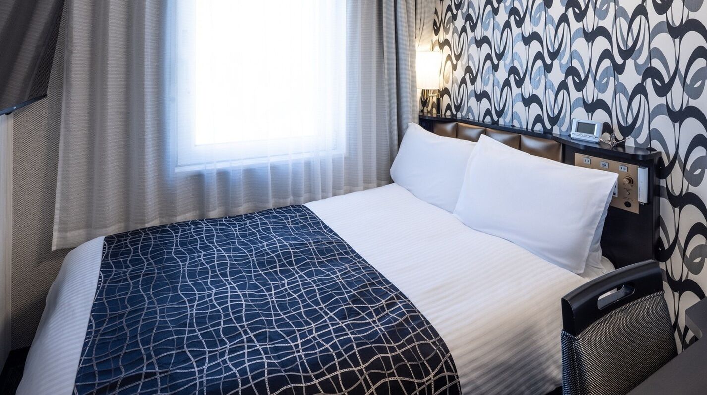 Een hotelkamer met een bed, hoofdbord, nachtkastje, lamp en een raam met gordijnen.