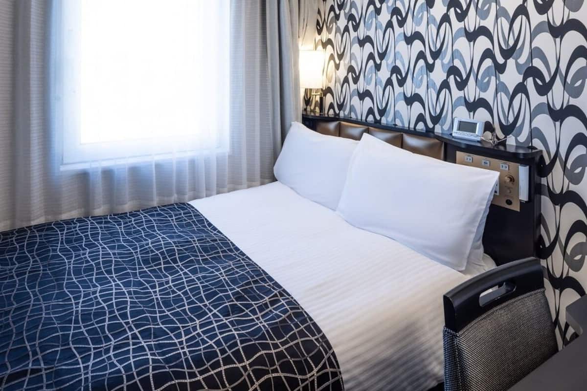 Habitación de hotel con cama, cabecera, mesita de noche, lámpara y ventana con cortinas.