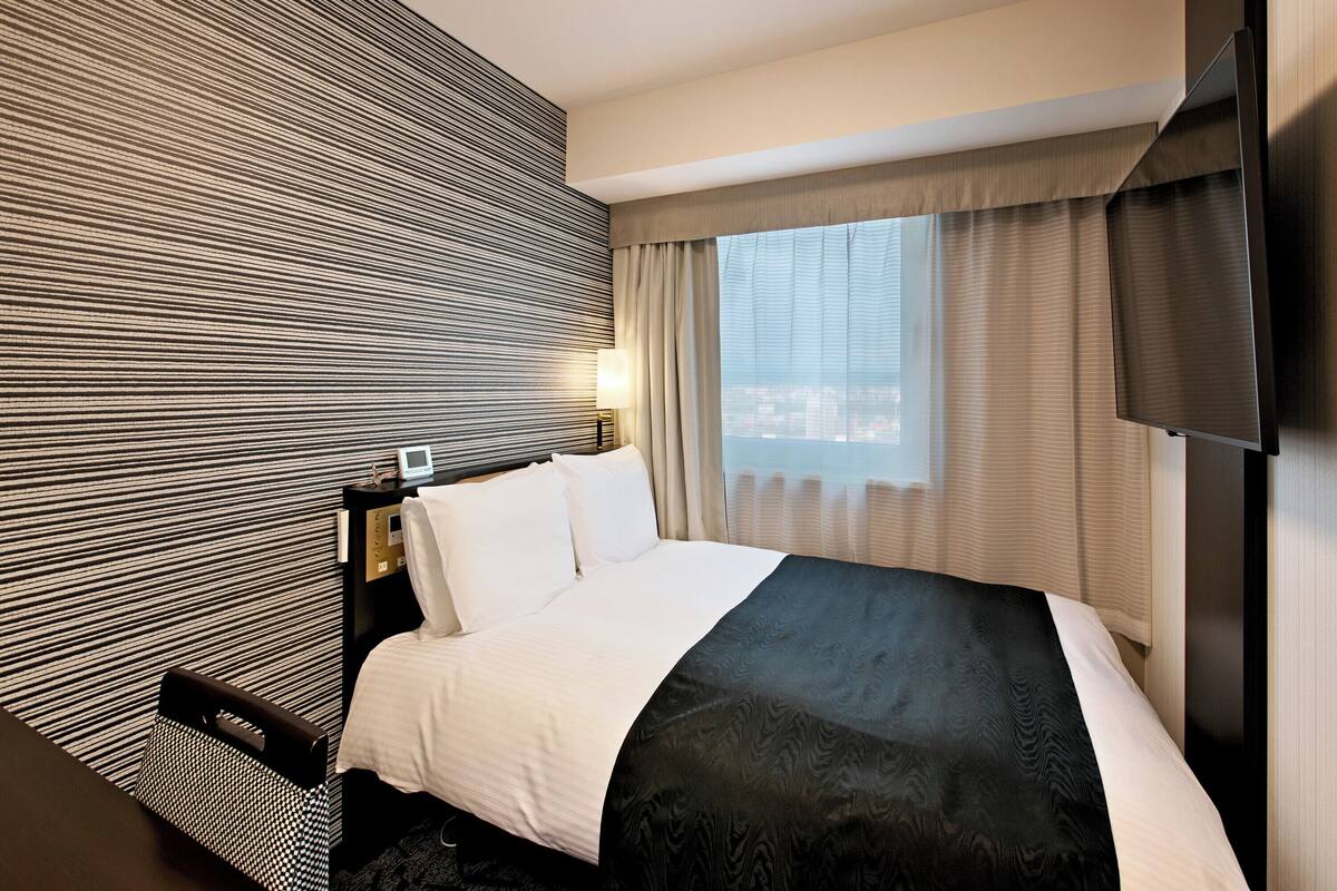 Habitación de hotel con una cama grande, un televisor de pantalla plana, un ventanal con cortinas y un papel tapiz estampado.