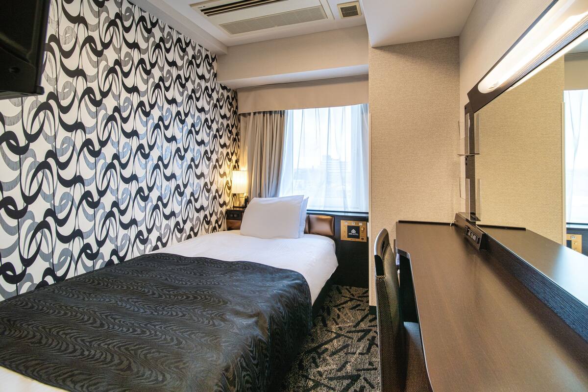 Una habitación de hotel con papel tapiz estampado, una cama con cubrecama oscuro, una mesita de noche con lámpara y un espejo grande.