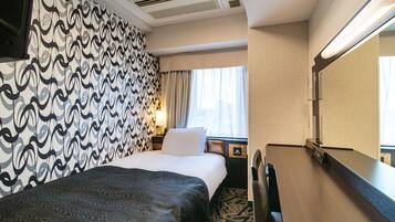 Une chambre d’hôtel avec un papier peint à motifs, un lit recouvert d’une couverture foncée, une table de chevet avec une lampe et un grand miroir.
