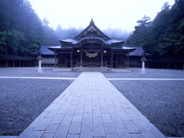Un templo japonés tradicional con un camino central que conduce al salón principal, rodeado por un patio de grava y un bosque denso.