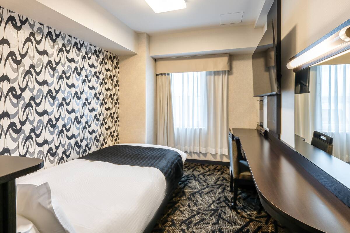 Una habitación de hotel con cama, escritorio, silla, televisión y ventana con cortinas.