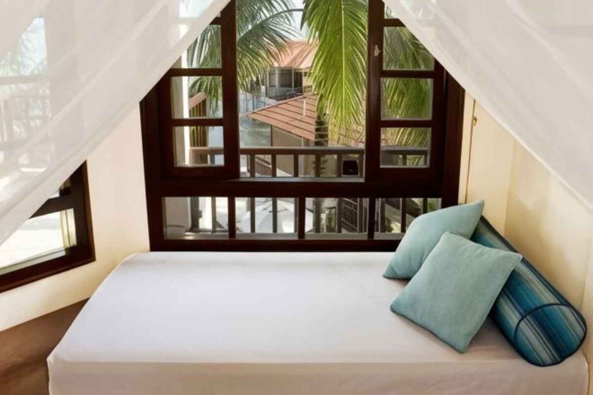 Una cama con colchón blanco y dos almohadas, una ventana con vistas a palmeras y un balcón con barandilla de madera.