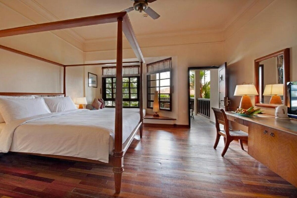 Una cama con dosel sobre un armazón de madera, un escritorio con lámpara, y vista al exterior a través de una ventana.