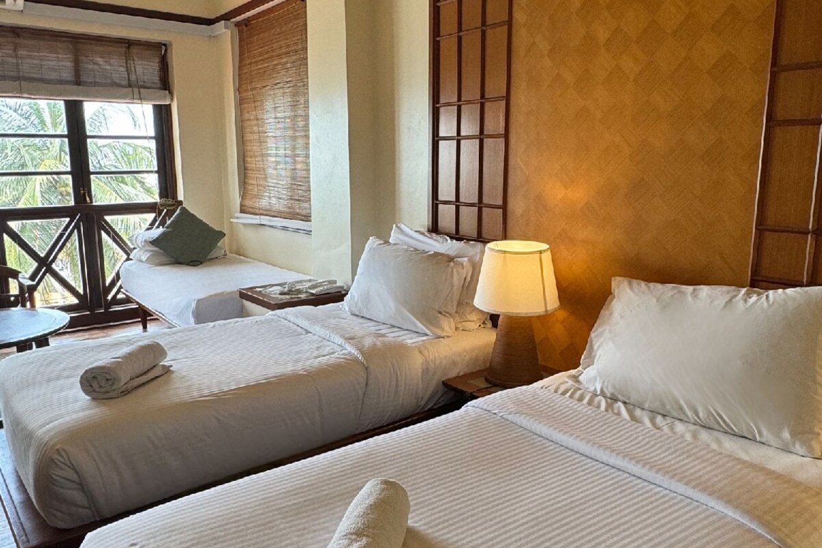Habitación de hotel con dos camas, una mesita de noche con lámpara, una ventana con persianas y vistas a palmeras.