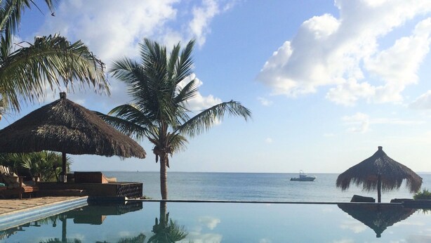 A clear infinity pool reflecting palm trees and a boat on the water. - Villas do Indico Eco - Resort & Spa Lodge (Vilanculos)