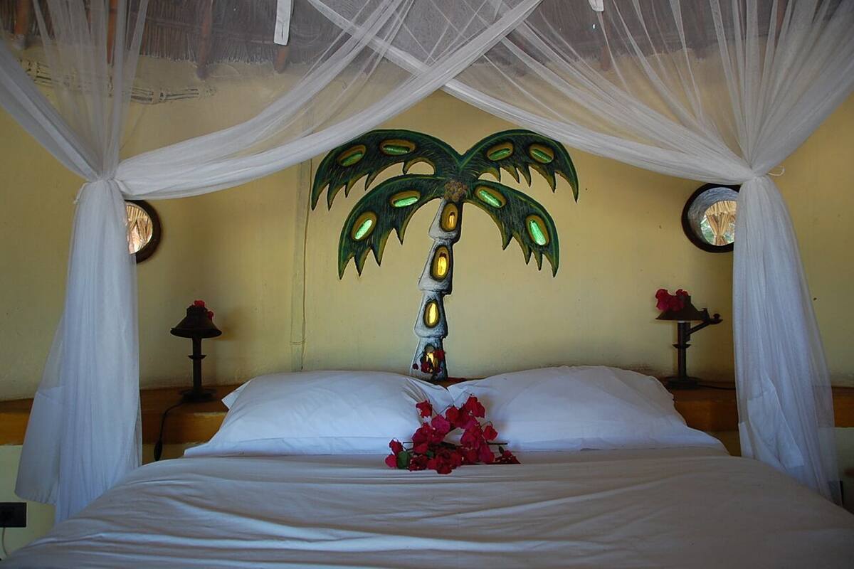 Ein Bett mit weißer Bettwäsche, eine dekorative Wandmalerei mit einer Palme und ein Strauß roter Blumen.