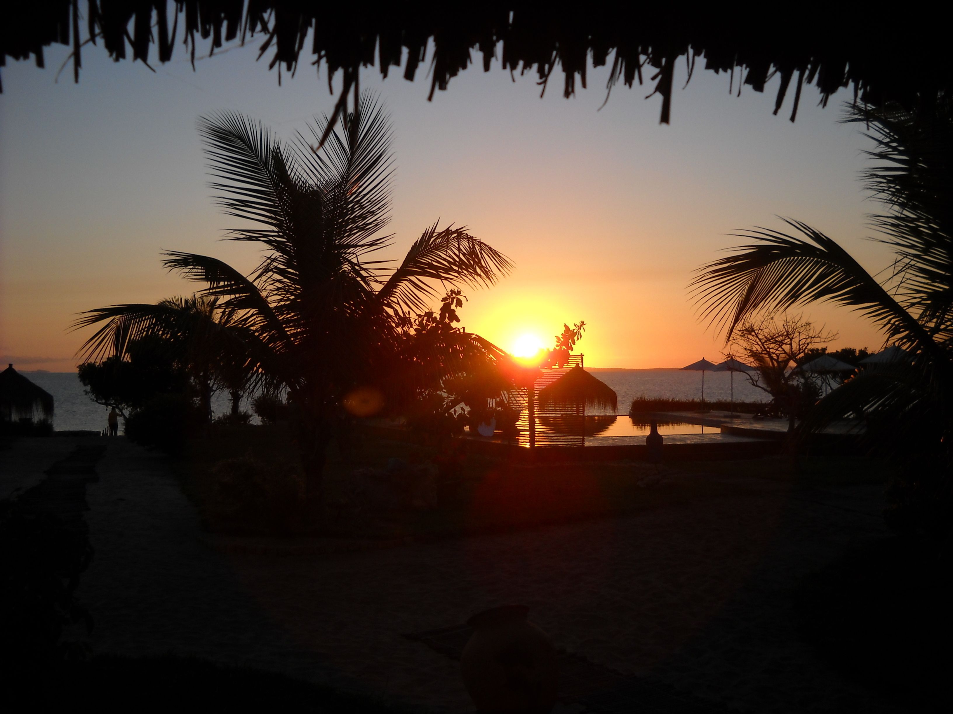 Tramonto su una spiaggia con palme e una costruzione a tetto di paglia.