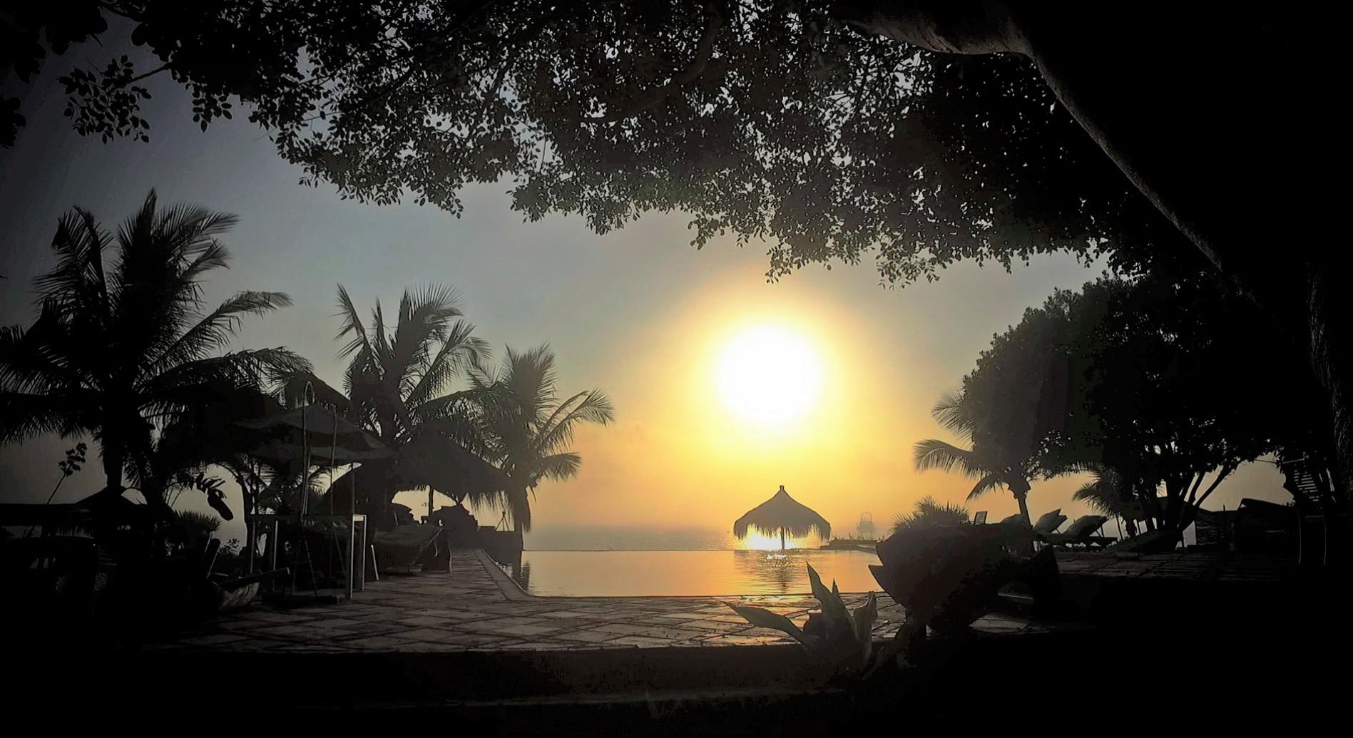 Escena de playa tropical con un atardecer, palmeras y una cabaña con techo de paja junto al agua.