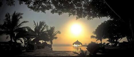 Escena de playa tropical con un atardecer, palmeras y una cabaña con techo de paja junto al agua.