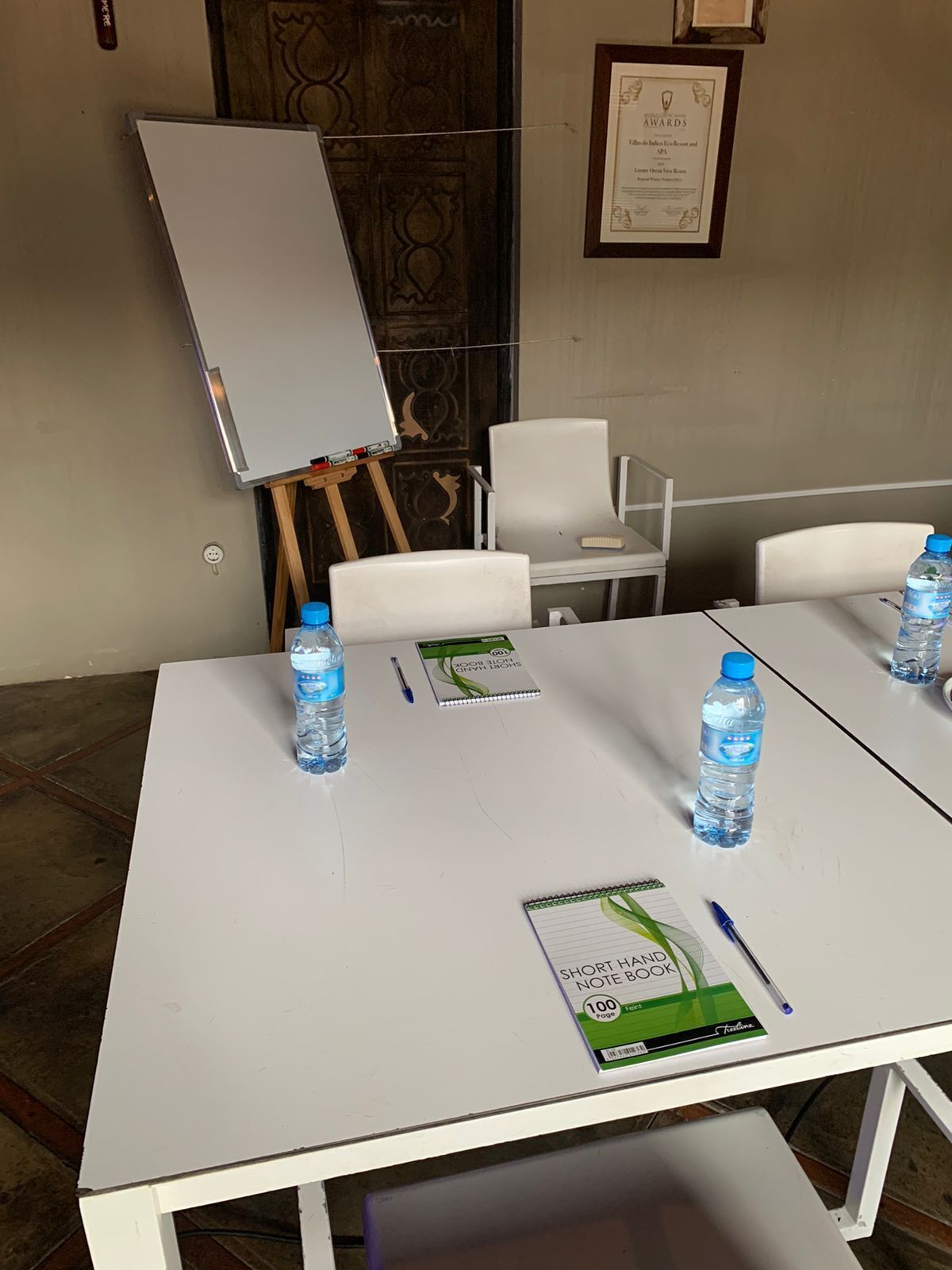 Une salle de conférence avec des tables et des chaises blanches, un tableau blanc et des bouteilles d’eau.