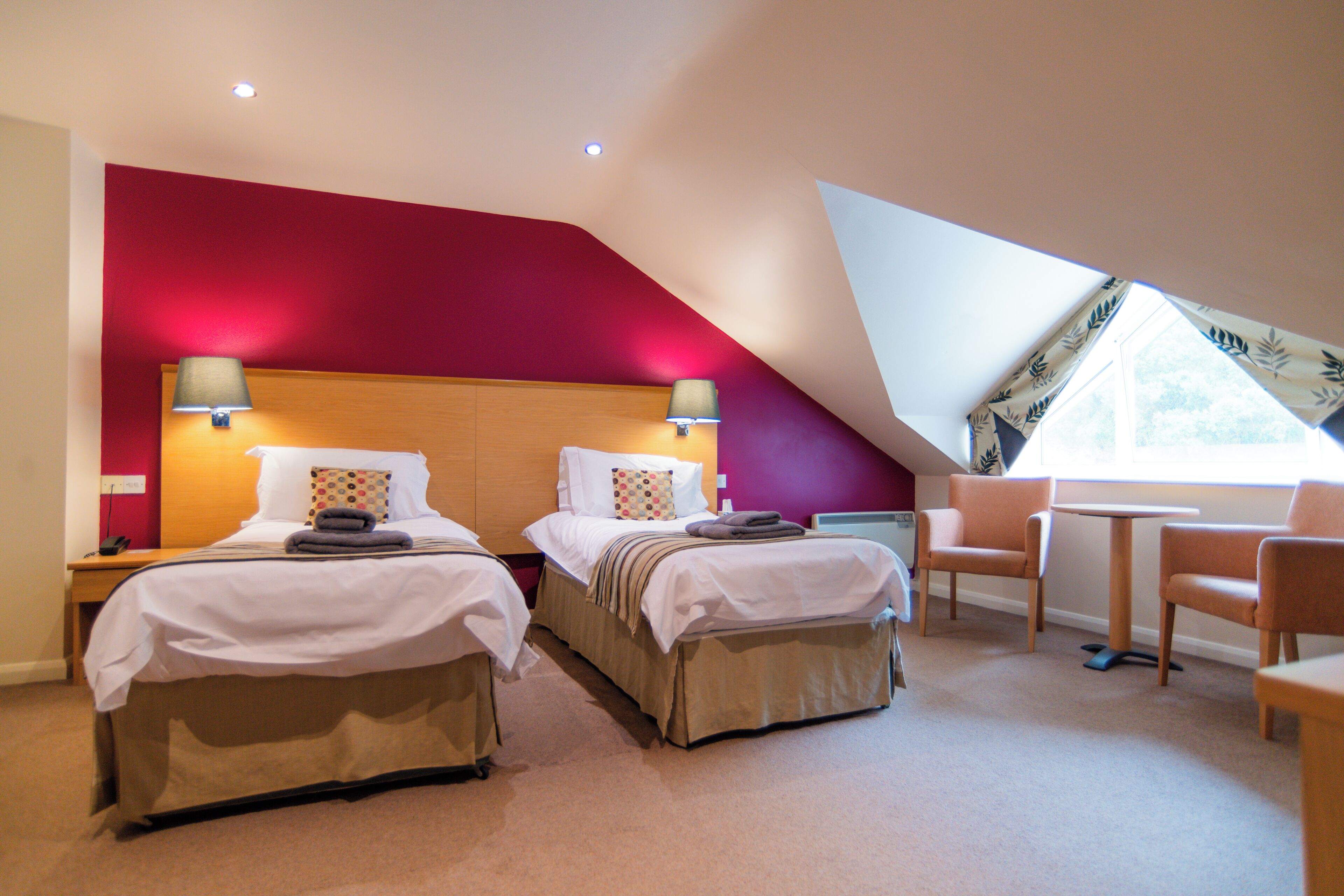 A hotel room with two beds, a red accent wall, a small table, and a chair.
