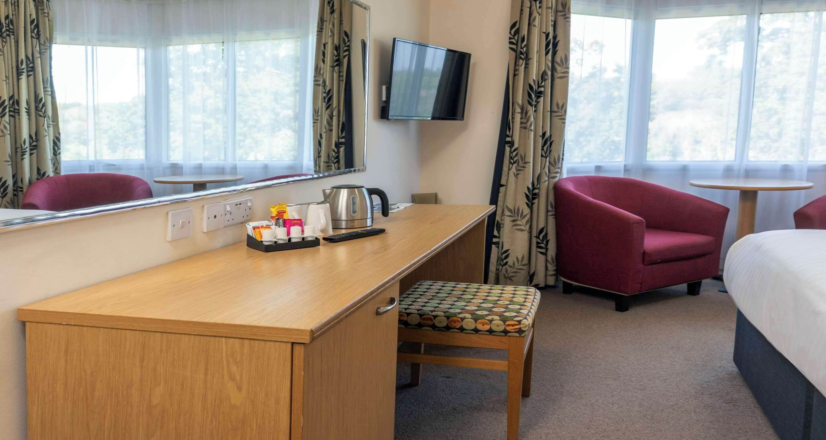 A hotel room with a wooden desk, a red armchair, a small table, a bed, and a television.
