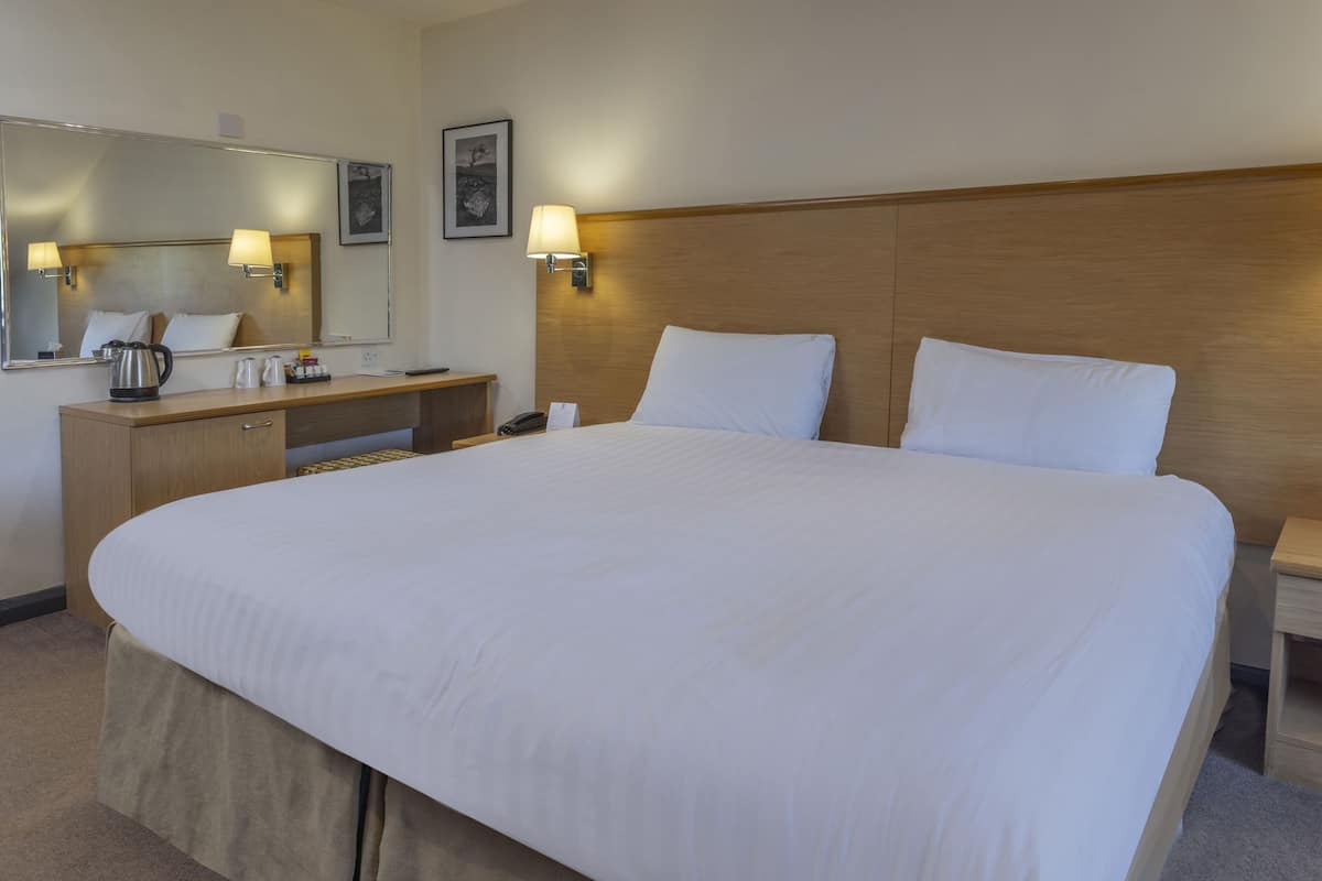 Una habitación de hotel con una cama grande, un cabecero de madera, una mesita de noche, un espejo y lámparas fijadas a la pared.