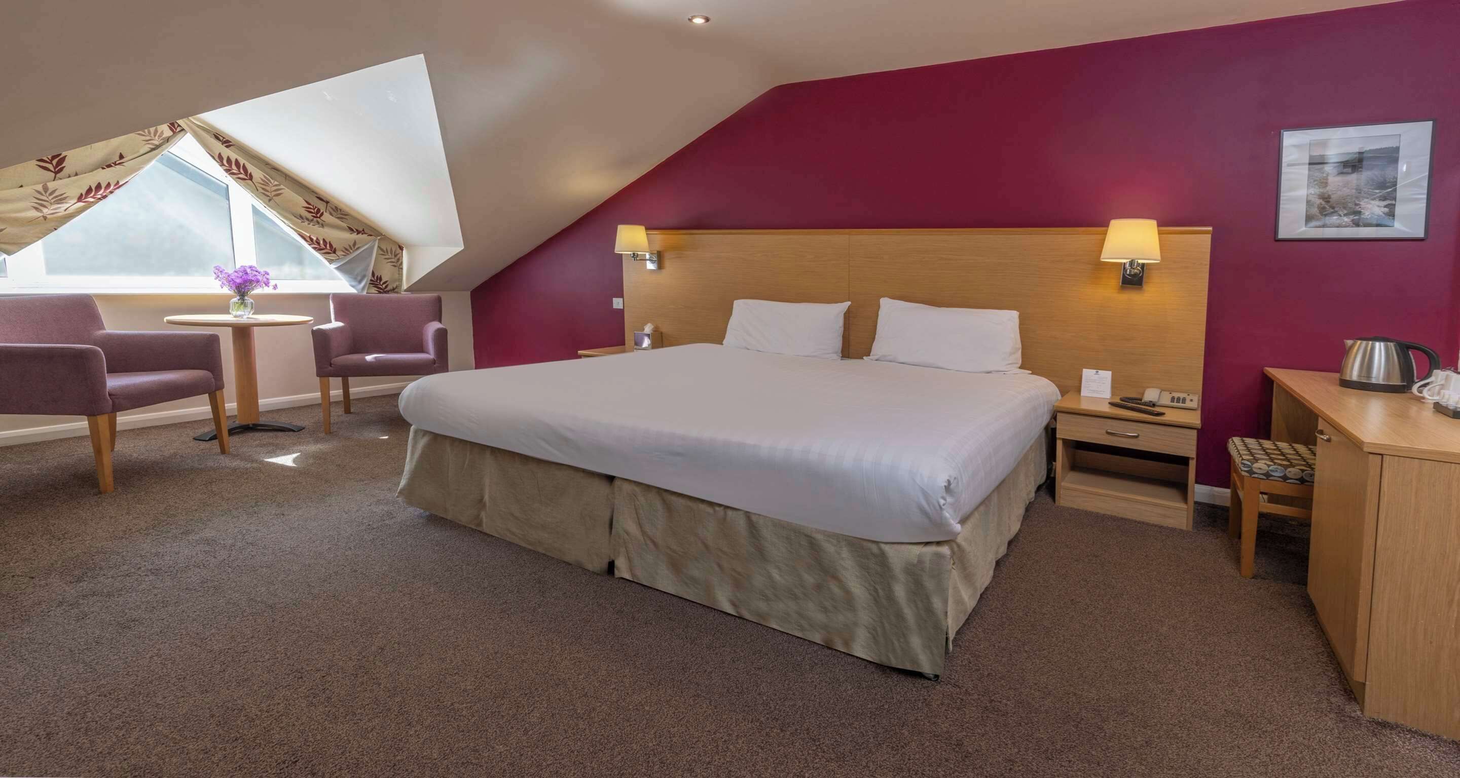 A hotel room with a bed, a desk, a chair, a small table with a vase of flowers, and a window with curtains.