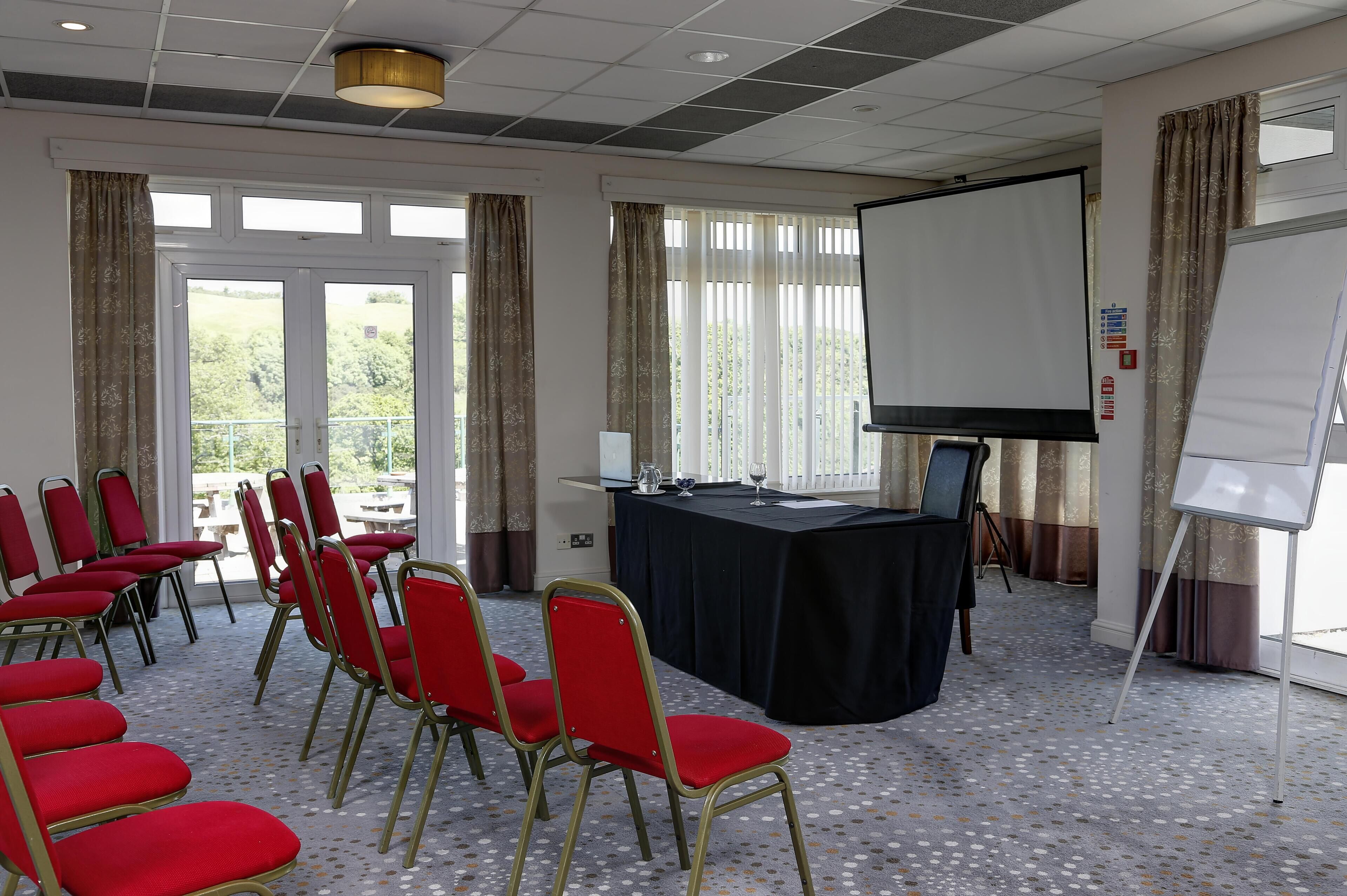 Une salle de conférence avec des chaises rouges disposées en rangées, une table noire sur laquelle est installé un écran de projecteur, et de grandes fenêtres avec des rideaux.