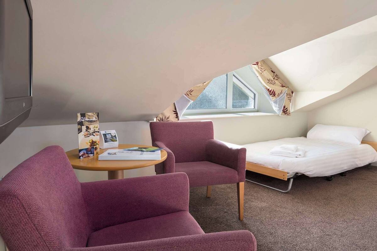 Una habitación pequeña y ordenada con una cama, un televisor, dos sillas, una mesa con un folleto y una ventana con cortinas.