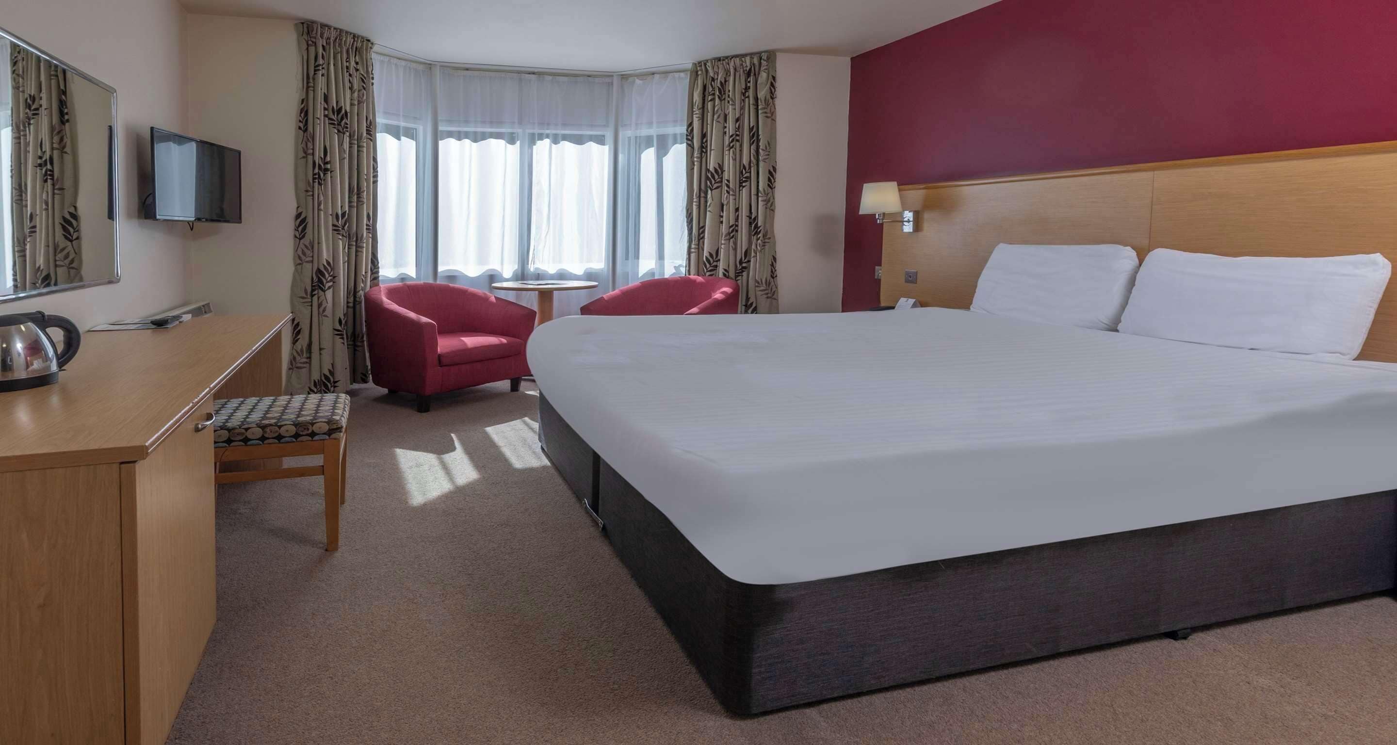 A hotel room with a large bed, a desk with a kettle, a chair, and a TV.
