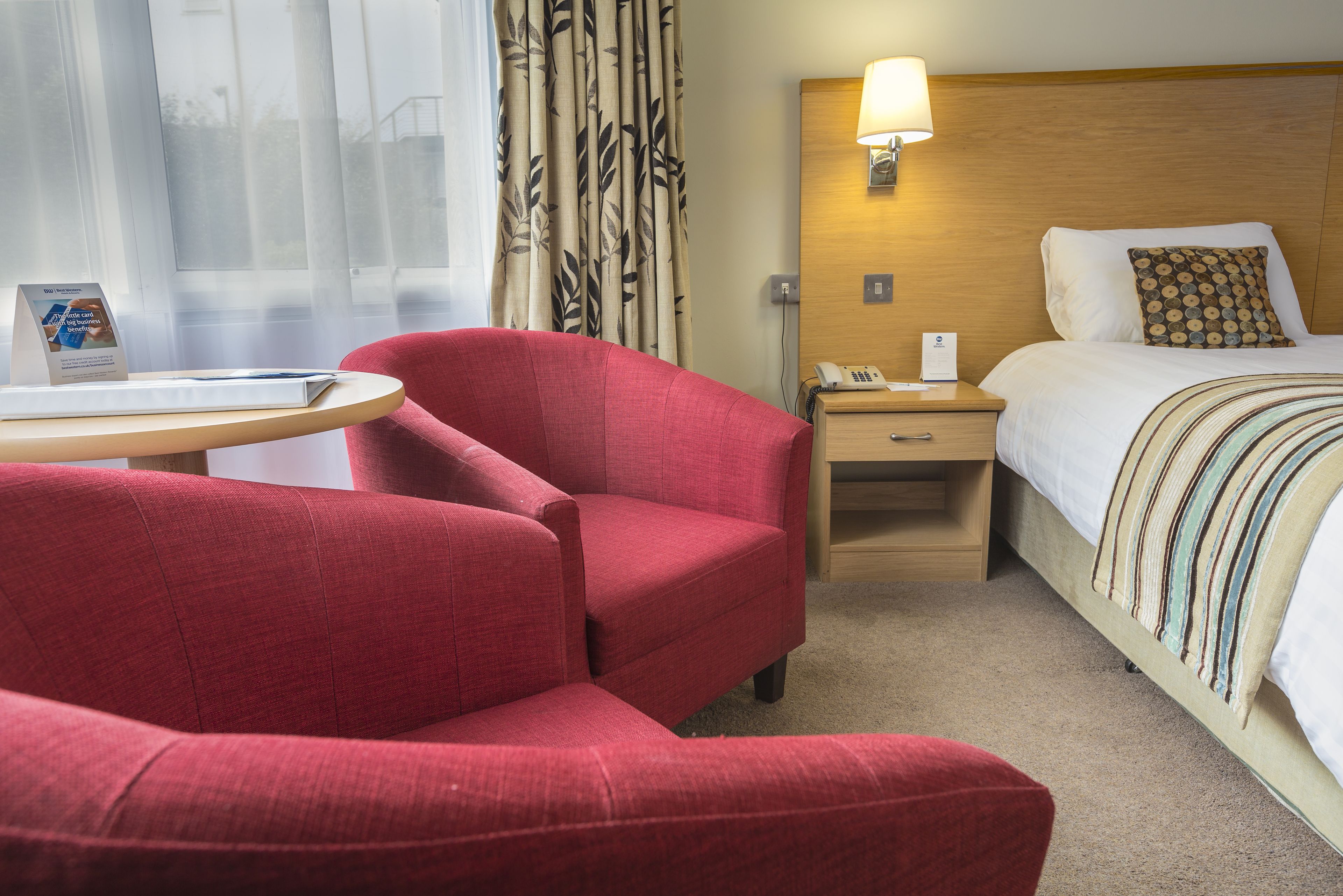 A hotel room with a red armchair, a bed with a striped blanket, a wooden headboard, a nightstand with a lamp, and a window with sheer curtains.
