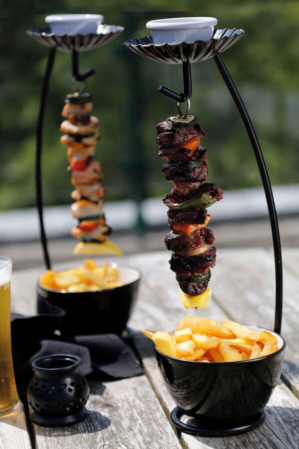 Dos brochetas de carne a la parrilla sobre una mesa de madera, con un bol de papas fritas y una pequeña vela.