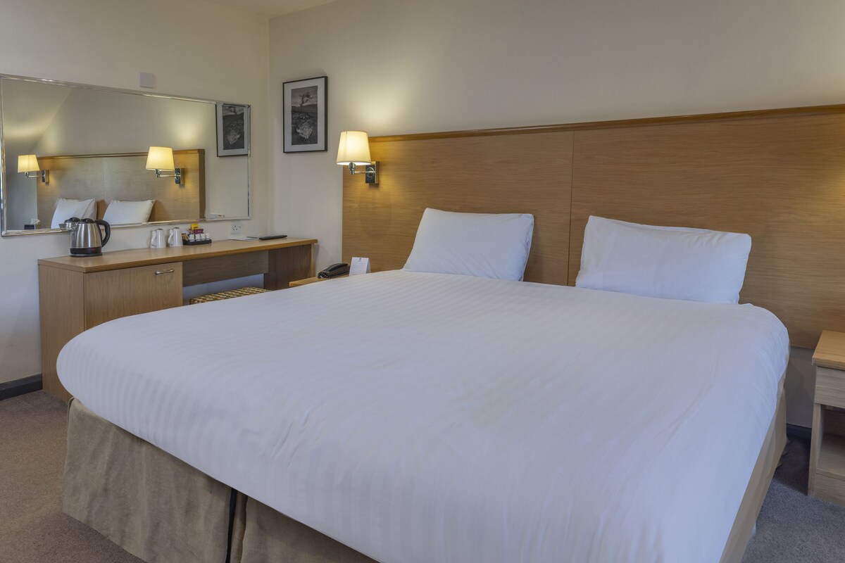 A hotel room with a large bed, a wooden headboard, a bedside table, a nightstand, a mirror, and wall-mounted lamps.