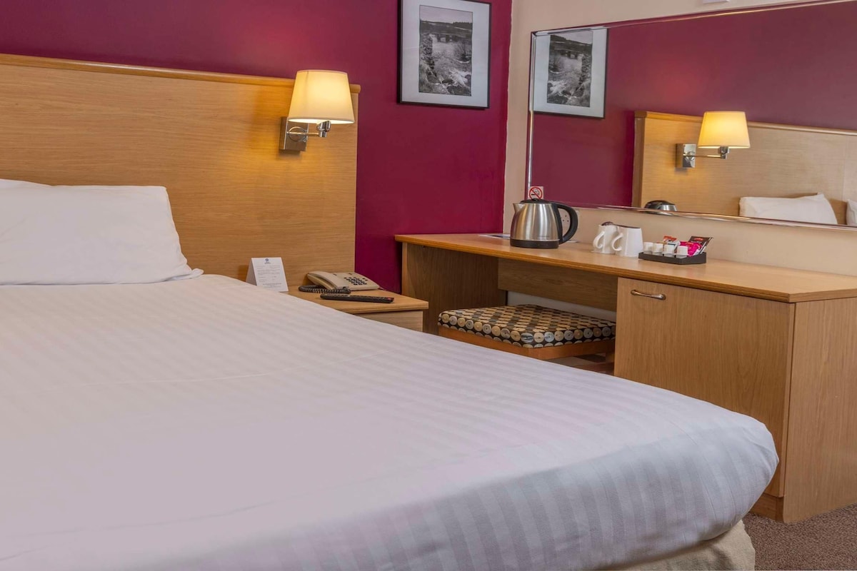 Habitación de hotel con una cama grande, lámparas de noche, un escritorio con un hervidor, un espejo y un taburete pequeño.