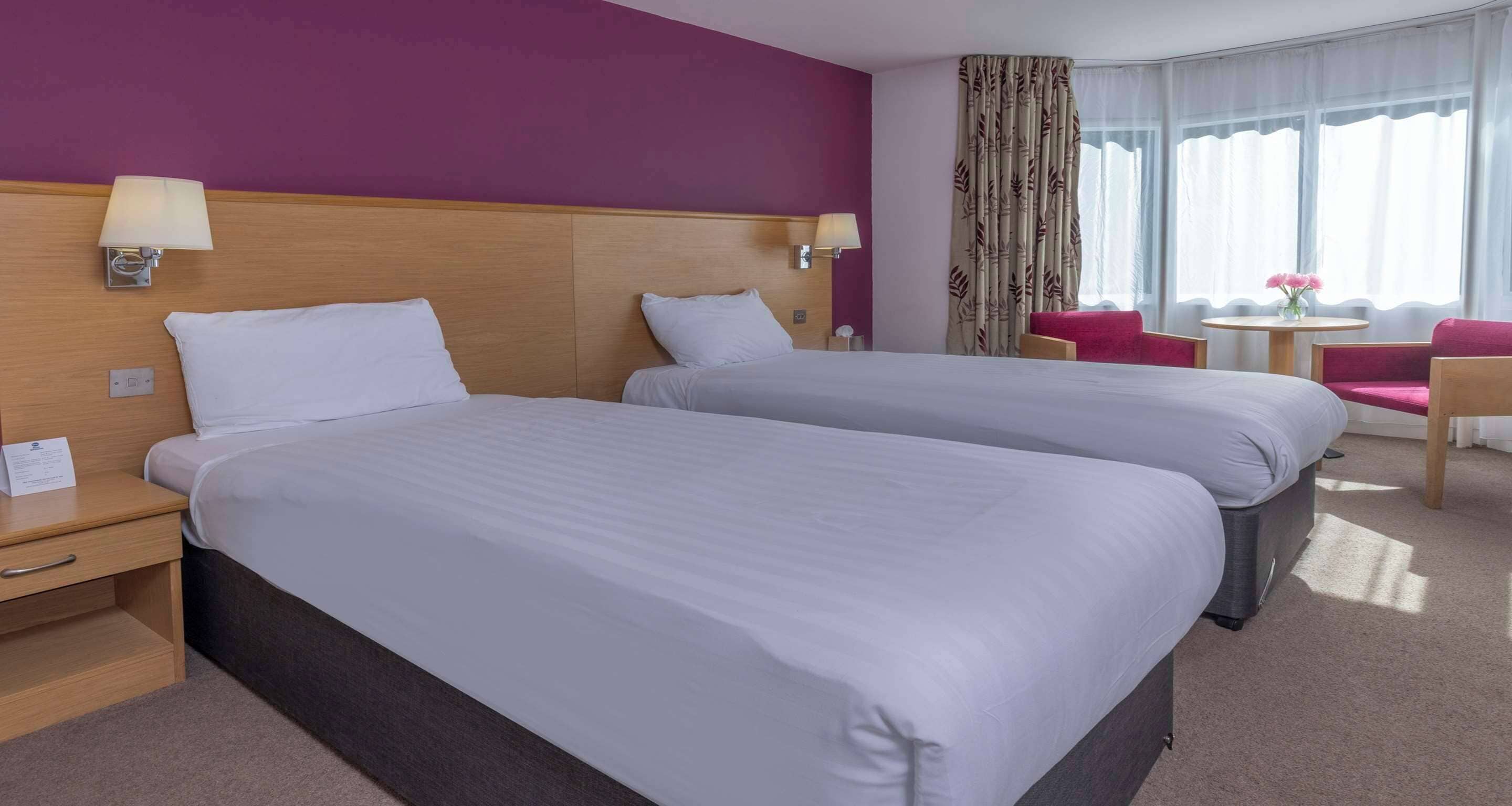 Habitación de hotel con dos camas, cabecero de madera, mesita de noche, escritorio, silla, pequeña mesa y ventana con cortinas.