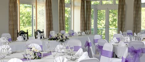 Een balzaal met gedekte tafels voor een formeel evenement, met witte tafellaken en paarse linten op de stoelen.