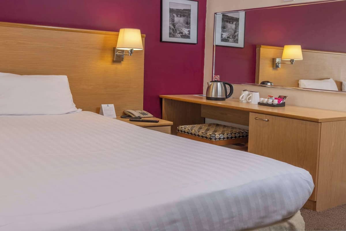 Una habitación de hotel con una cama grande, dos mesitas de noche, un escritorio con silla, un espejo y un pequeño rincón de descanso.