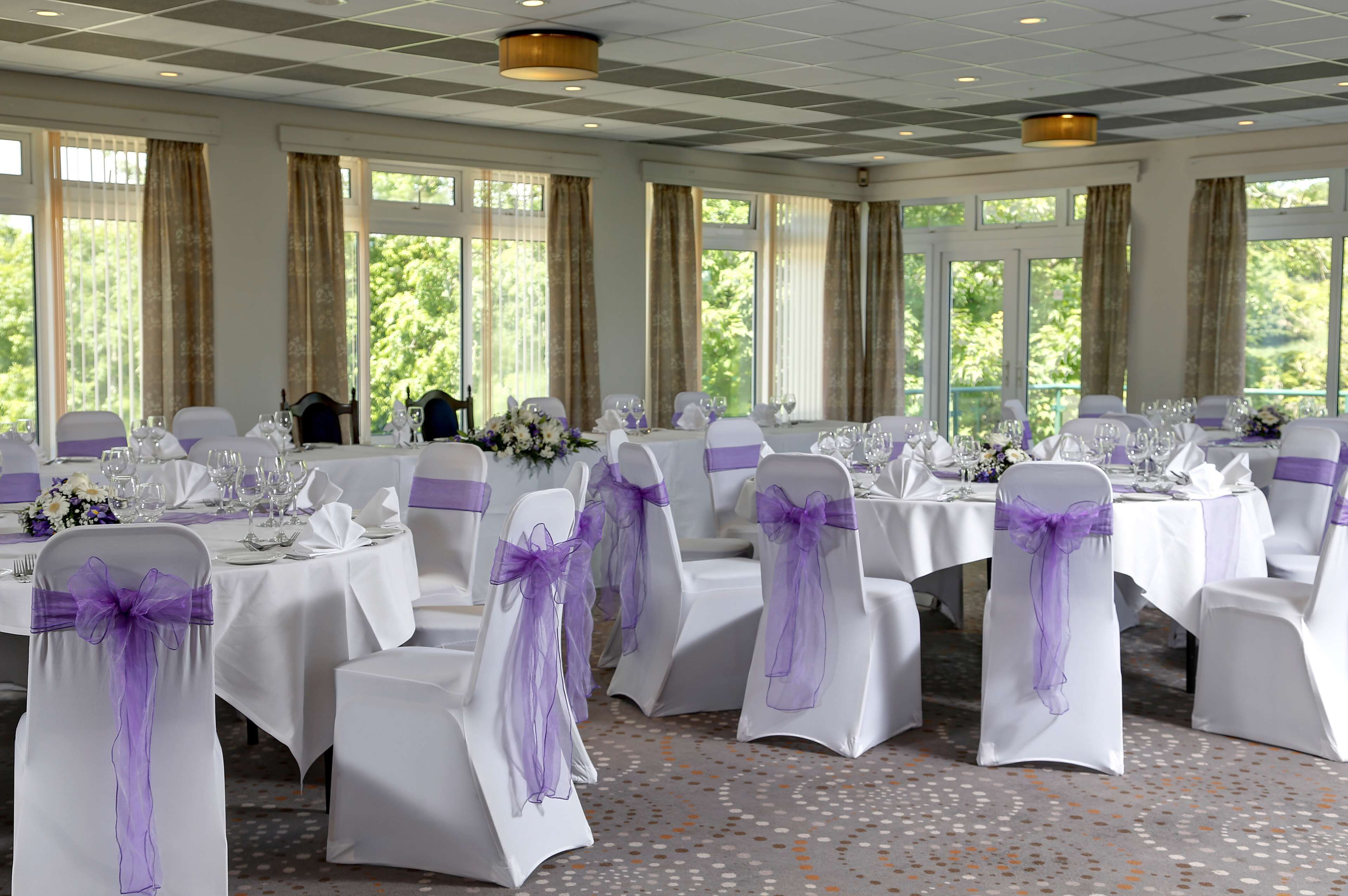 A banquet hall with white tables, purple sashes, and large windows.