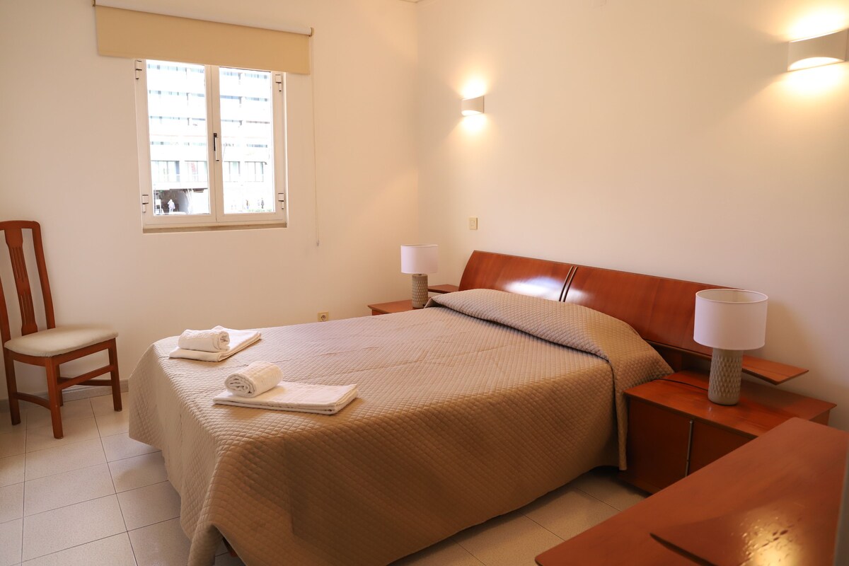 Une chambre d’hôtel avec un grand lit, une chaise en bois, deux tables de chevet avec des lampes et une fenêtre avec des stores.
