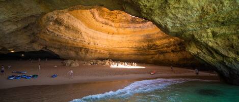 Una cueva con una gran formación rocosa y una zona de playa con gente y barcos.