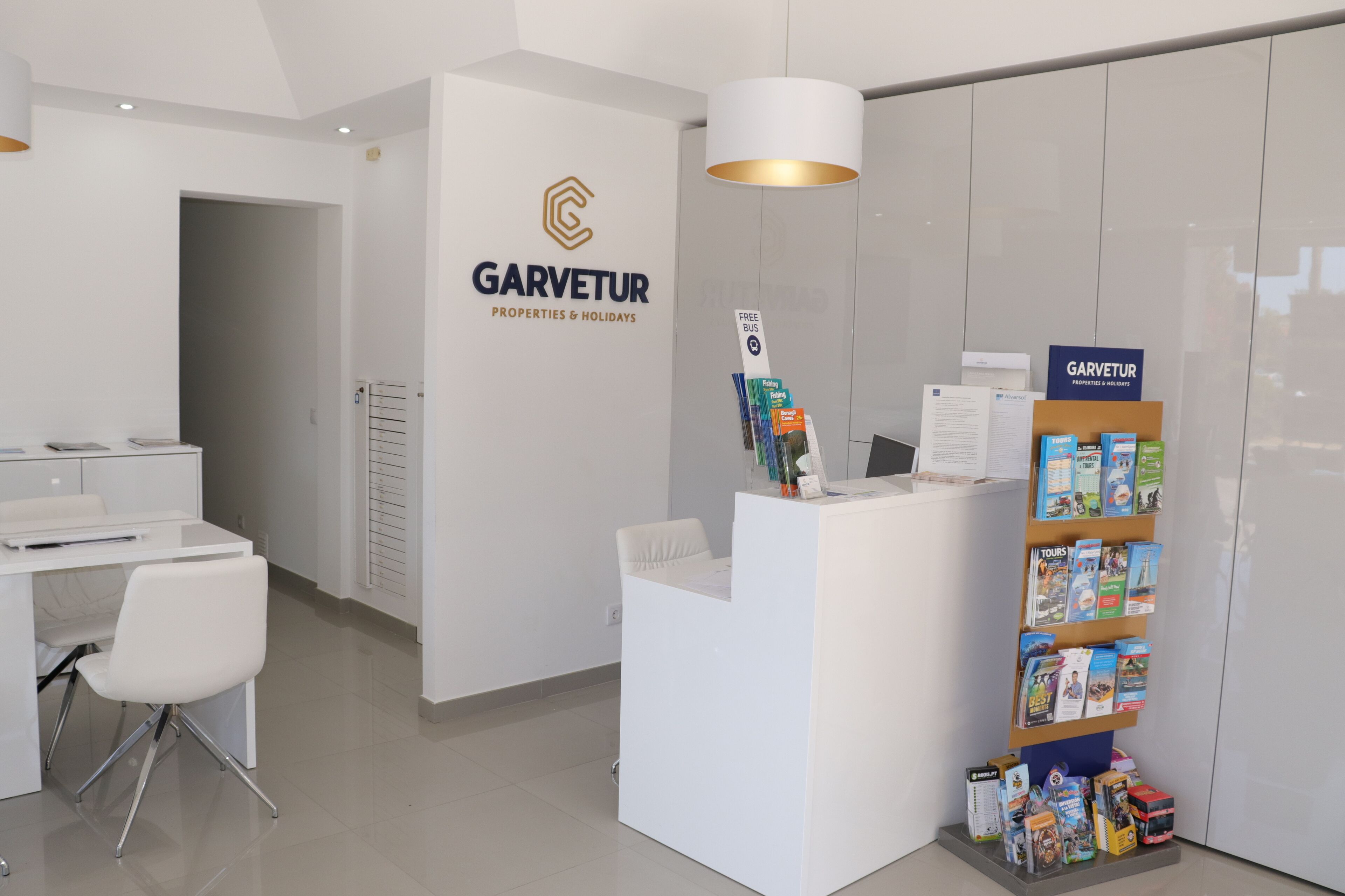 Un espace d’accueil comprenant un bureau blanc, une chaise, un comptoir orné d’un logo et un présentoir rempli de brochures de voyage.